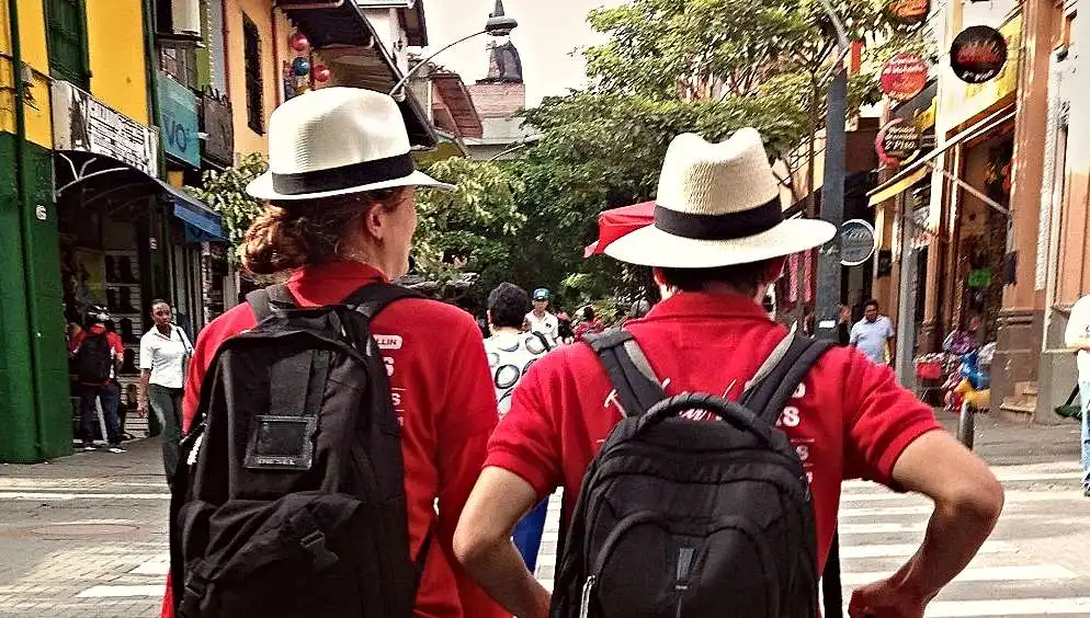 Two Real City Tours guides in red polo shirts and Panama hats walking down a busy Medellín commercial street
