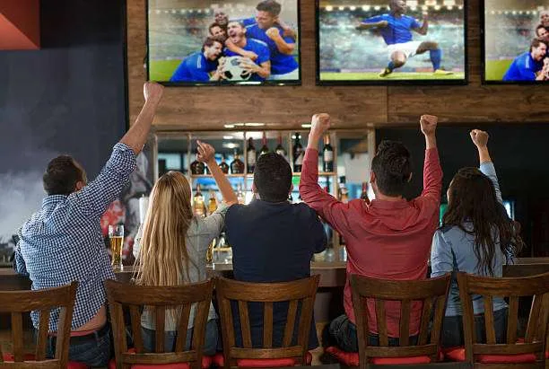 Group of friends cheering with fists raised at a sports bar watching football on multiple big screens