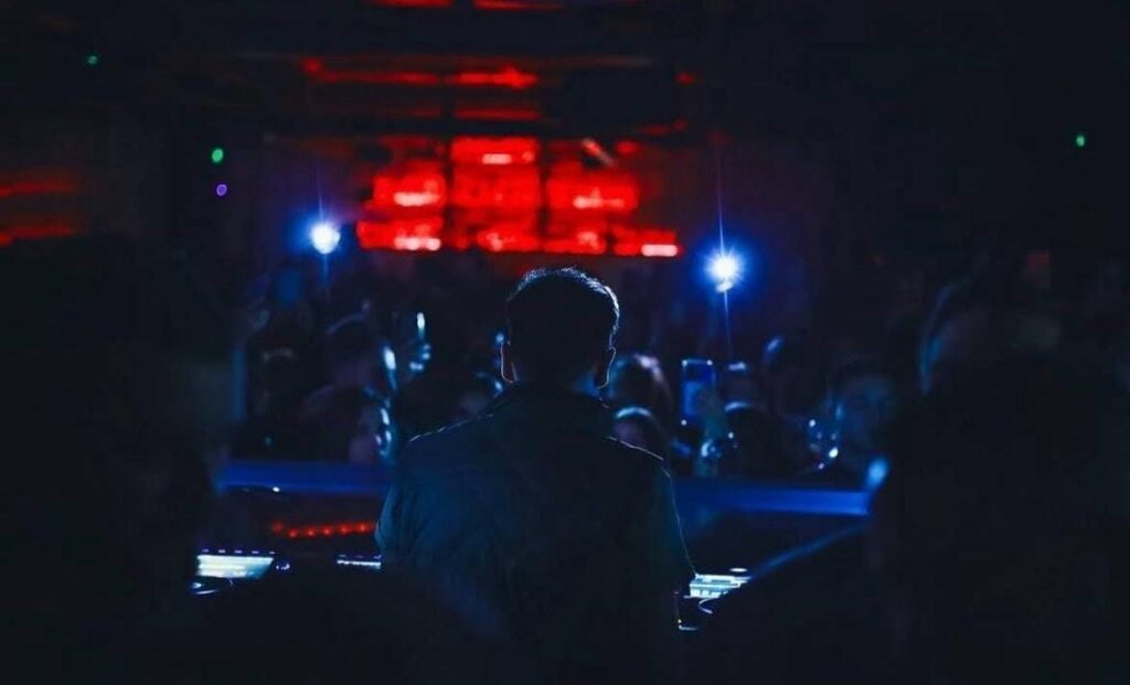 Silhouette of DJ at turntable facing dancing crowd with blue and red stage lighting in Medellín