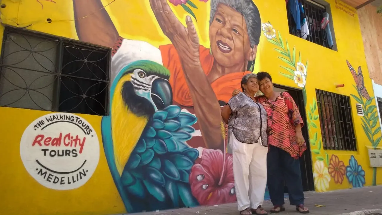 Two local women posing in front of a bright yellow mural with Real City Tours Medellín logo and parrot