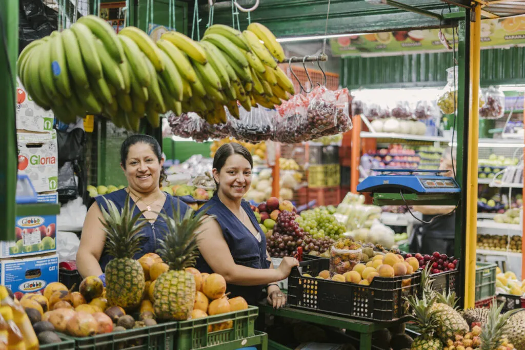 Two smiling women vendors at a fruit stall in Medellín market surrounded by pineapples and tropical fruits