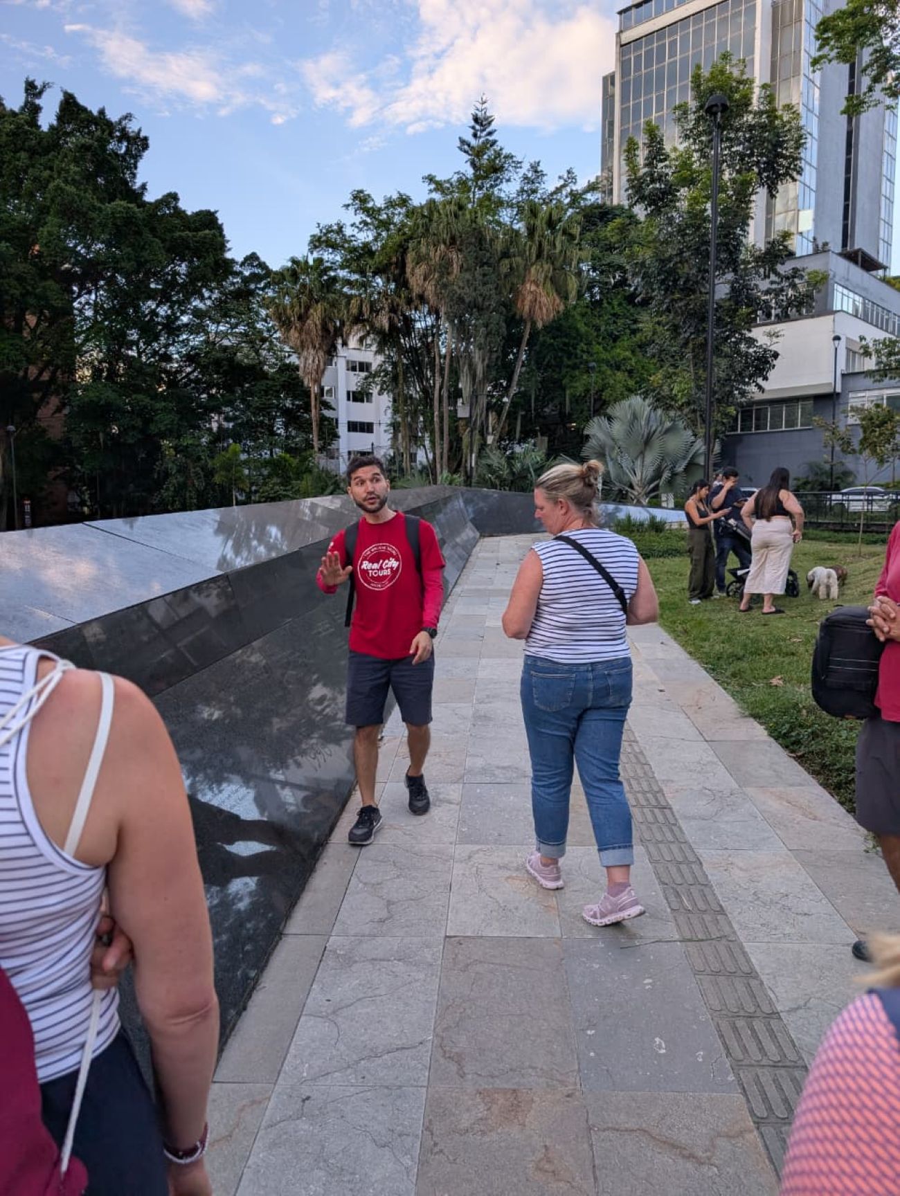 Real City Tours guide in red shirt speaking to tourists beside a dark marble memorial wall in Medellín