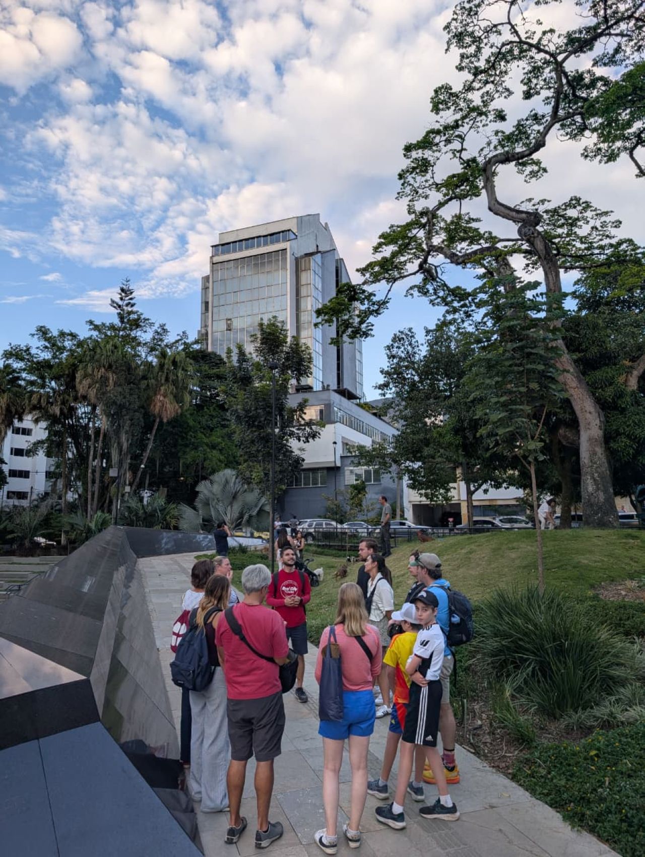 Real City Tours guide with tourists gathered around a dark angular memorial monument in a Medellín park