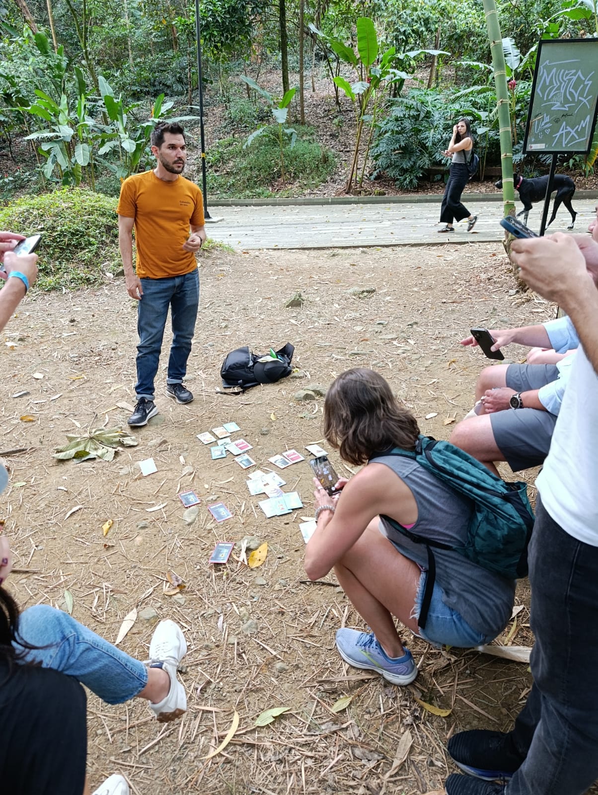 Tour guide in orange shirt presenting historical cards to tourists seated on the ground in a Medellín park